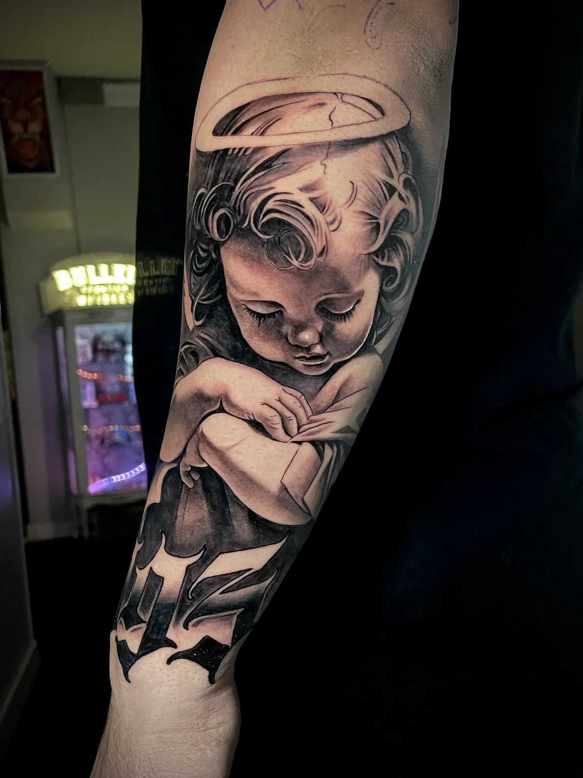 Black and grey realism cherub angel baby with halo and lettering forearm tattoo by Andras Tattoo